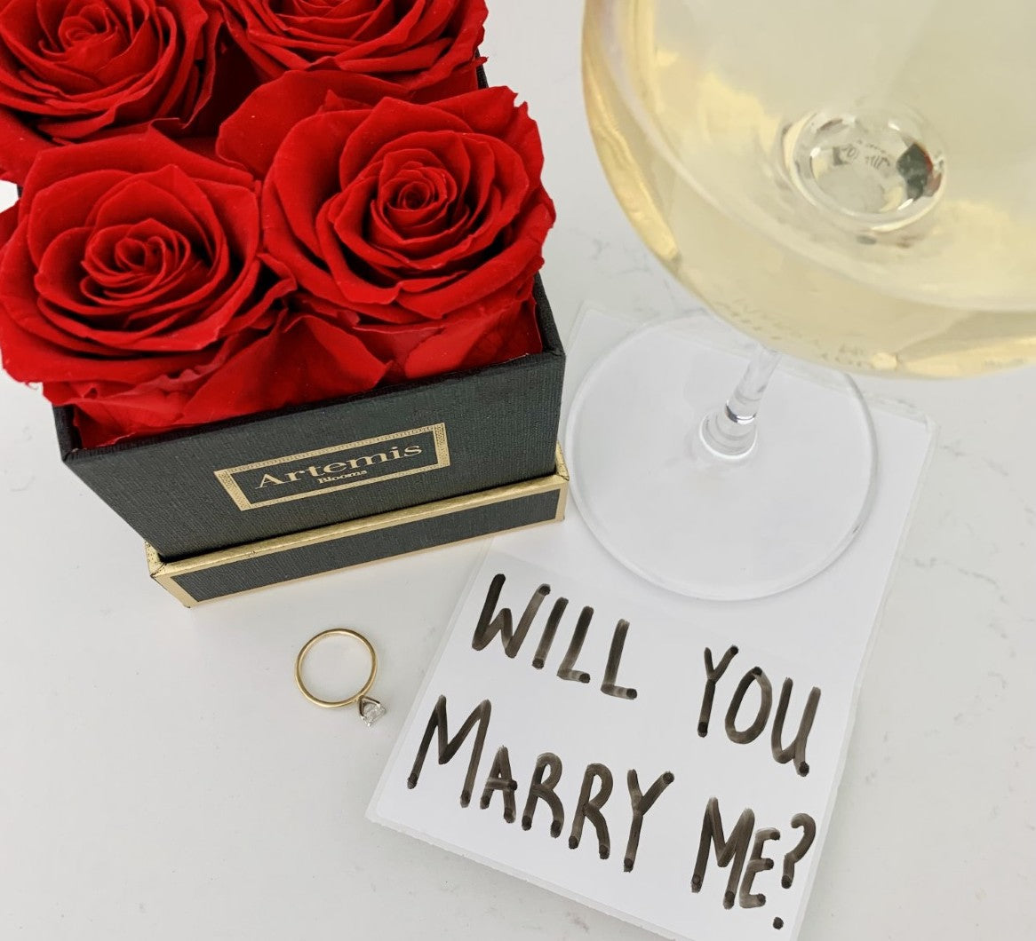 A marriage proposal with red roses in a black box with an engagement ring and white wine in a glass